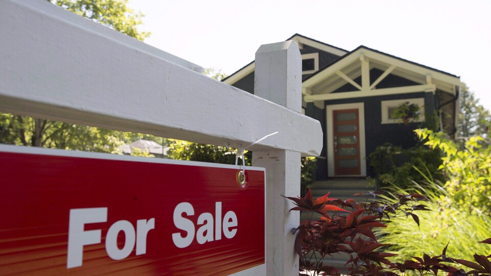 Une pancarte « À vendre », en anglais, en avant-plan de l'image devant une maison.