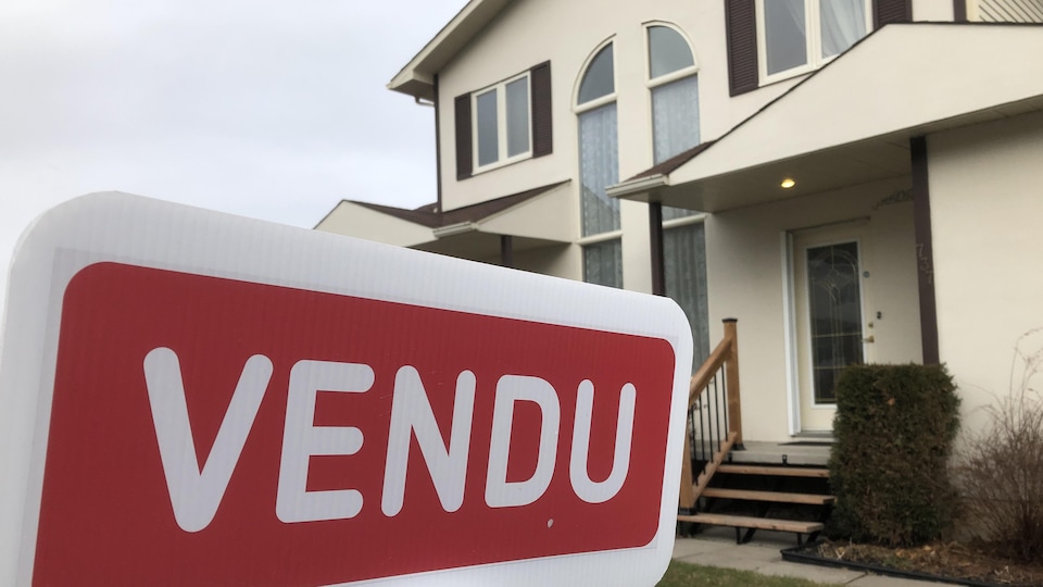 Une pancarte de maison indiquant Vendu, et à l'arrière-plan une maison de banlieue.