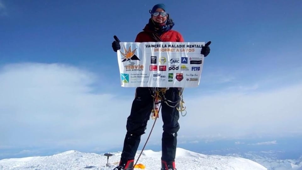 Jean-François Dupras, habillé en alpiniste, est au sommet d'une montagne. Il tient une banderole où est écrit Vaincre la maladie mentale un sommet à la fois. 
