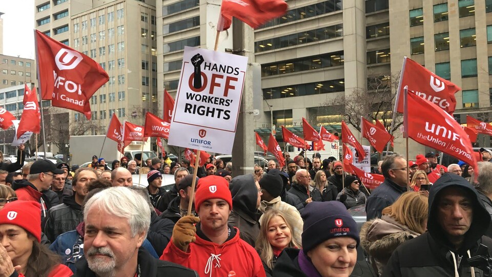 Des manifestants entre des édifices du centre-ville de Toronto tiennent haut dans les airs des drapeaux avec le symbole d'Unifor et des slogans syndicaux sur ceux-ci.