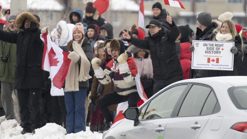 Des manifestants avec des drapeaux du Canada et des pancartes applaudissent les voitures qui passent.
