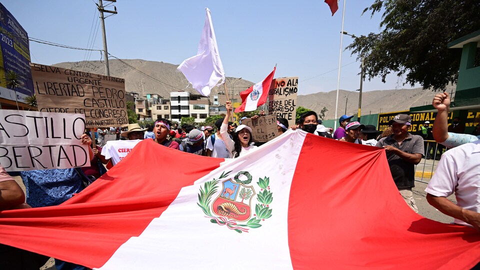 Des hommes et des femmes tiennent dans leurs mains un immense drapeau du Pérou ainsi que des pancartes avec des slogans dans une rue de Lima. 