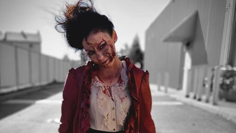 Une femme costumée en zombie portant une jupe noire, une veste rouge, les cheveux en bataille et des traces de sang sur le visage. 