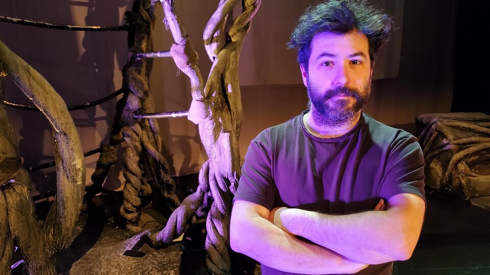 Marc-André Charron, les bras croisés, en face d'un décor de théâtre représentant un arbre. 