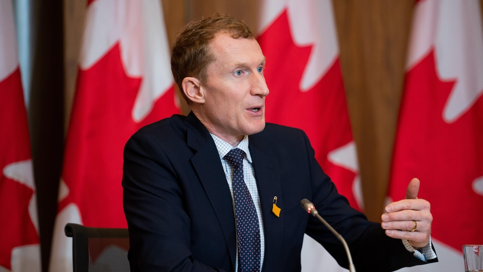 Le ministre parle assis à une table lors d'une conférence de presse avec des drapeaux du Canada en toile de fond.