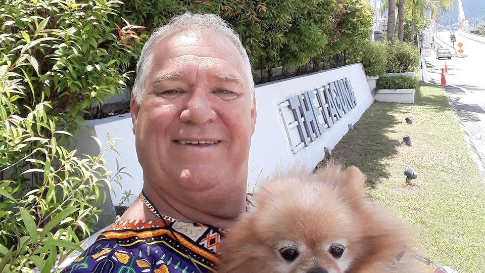Égoportrait d'un homme avec un chien dans ses bras devant un décor tropical.