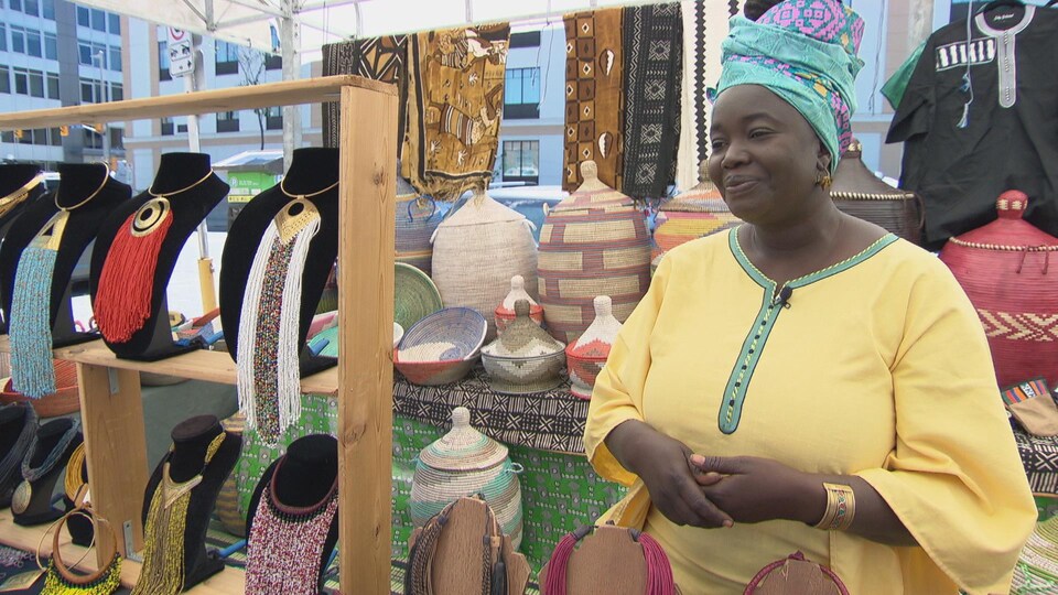 La joaillière d'origine sénégalaise est debout, dans son kiosque, entourée des colliers et des paniers tressés qu'elle y vend.