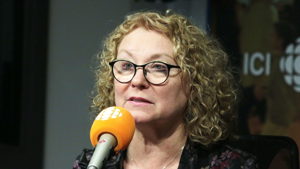 Une femme aux cheveux blonds bouclés et portant des lunettes de vue à cadre noir parle au micro.