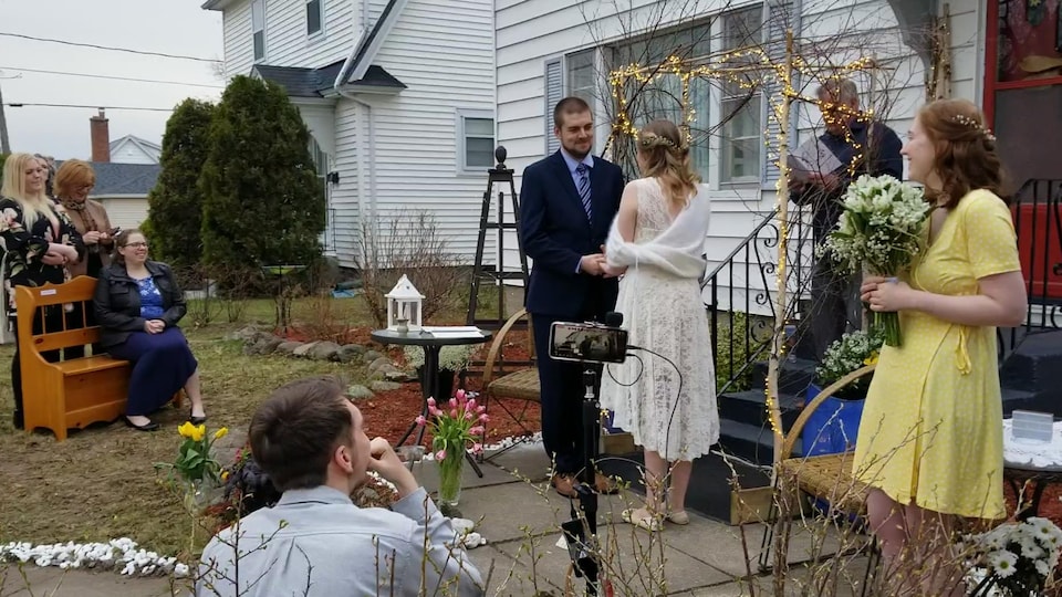 Un couple entouré de quelques personnes est debout devant une maison et échange leurs voeux de mariage.
