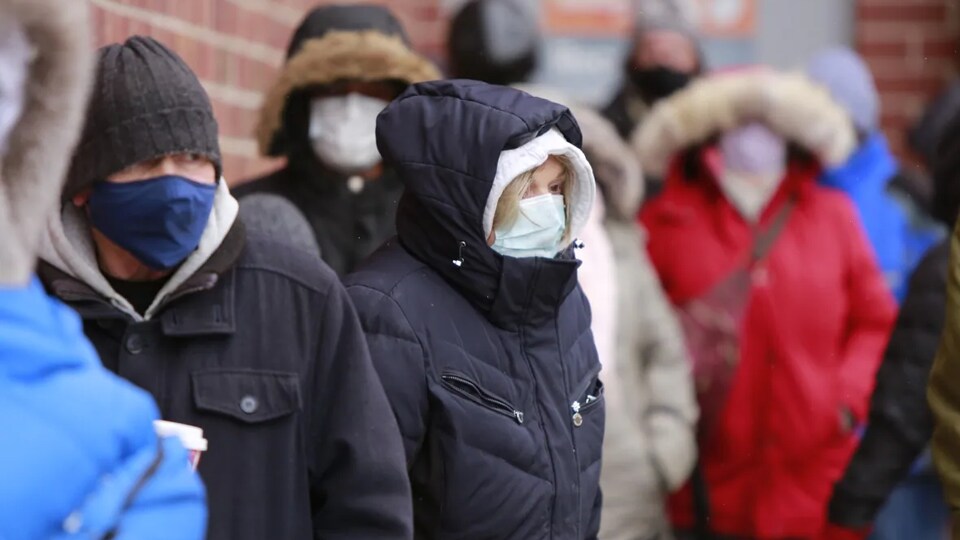 Des gens bien emmitouflés dans leur manteau d’hiver, et portant des masques dans une file d’attente à côté d’un bâtiment en brique.