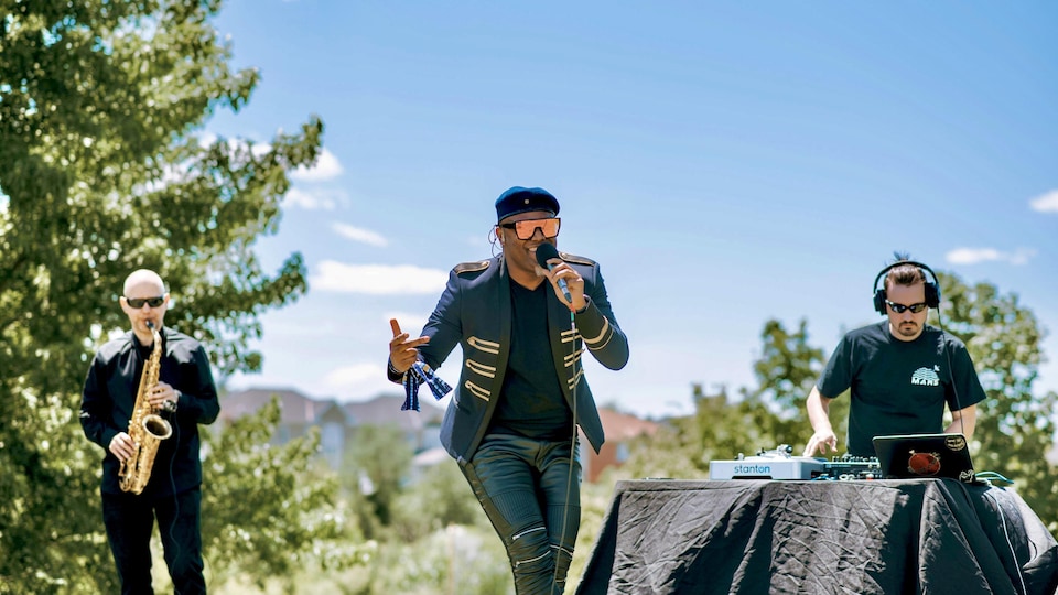 Abel, le micro en main, sur une scène en plein air avec deux de ses musiciens.