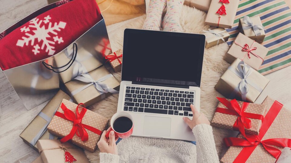 Un ordinateur portable sur les genoux d'une personne assise sur un tapis, avec de nombreux cadeaux pour les fêtes tout autour.