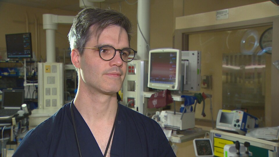 Le Dr Samuel Vaillancourt porte une blouse bleue marine, il a des lunettes, les cheveux poivre et sel, il est dans la salle de traumatologie