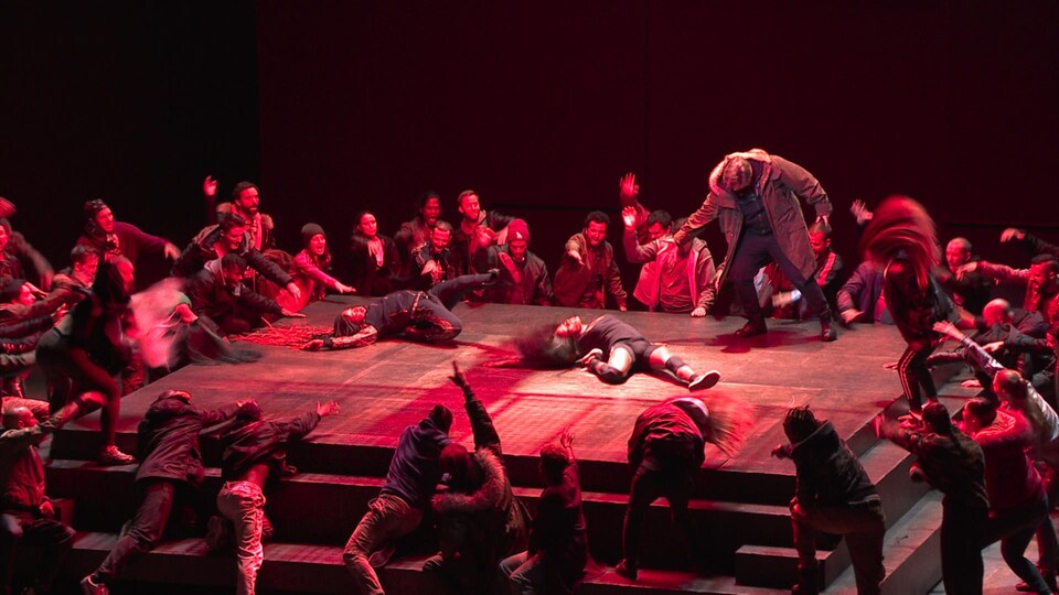 Une scène est éclairée en rouge, deux danseurs sont allongés, entourés par d'autres danseurs figurant une foule en colère.