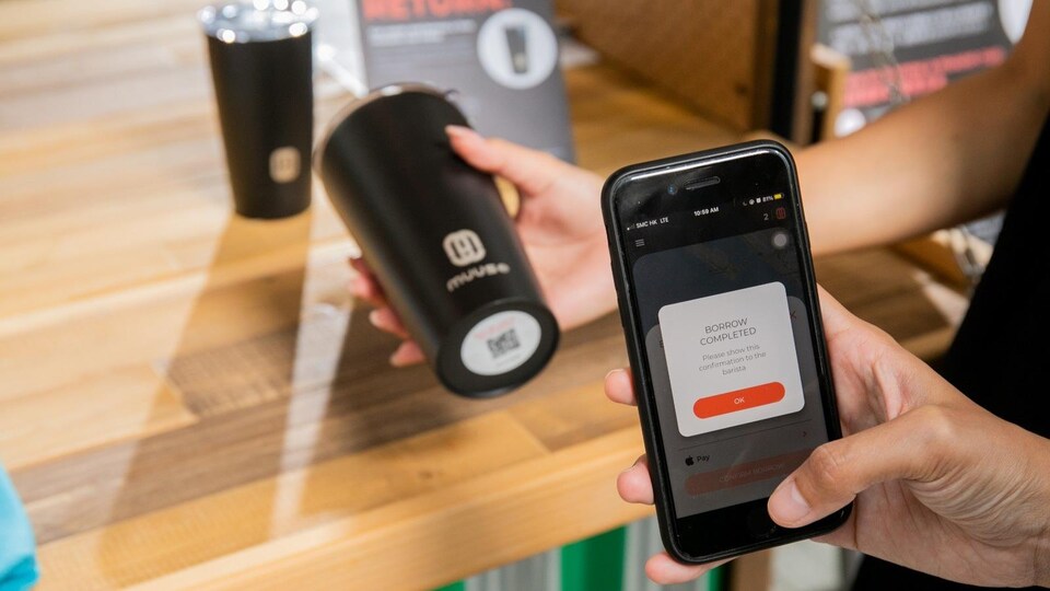 Une personne utilise un téléphone intelligent pour emprunter une tasse réutilisable. Des tasses réutilisables marquées « Muuse » se trouvent en arrière plan.