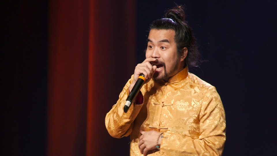 Anto Chan porte une veste chinoise jaune, ses cheveux sont attachés et il a un micro à la main.