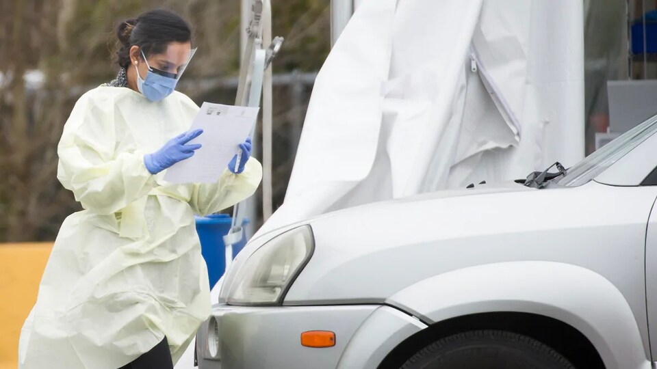 Une membre du personnel médical vêtue d'une jaquette et d'un masque lit un document devant une voiture à un centre de dépistage du coronavirus au volant.