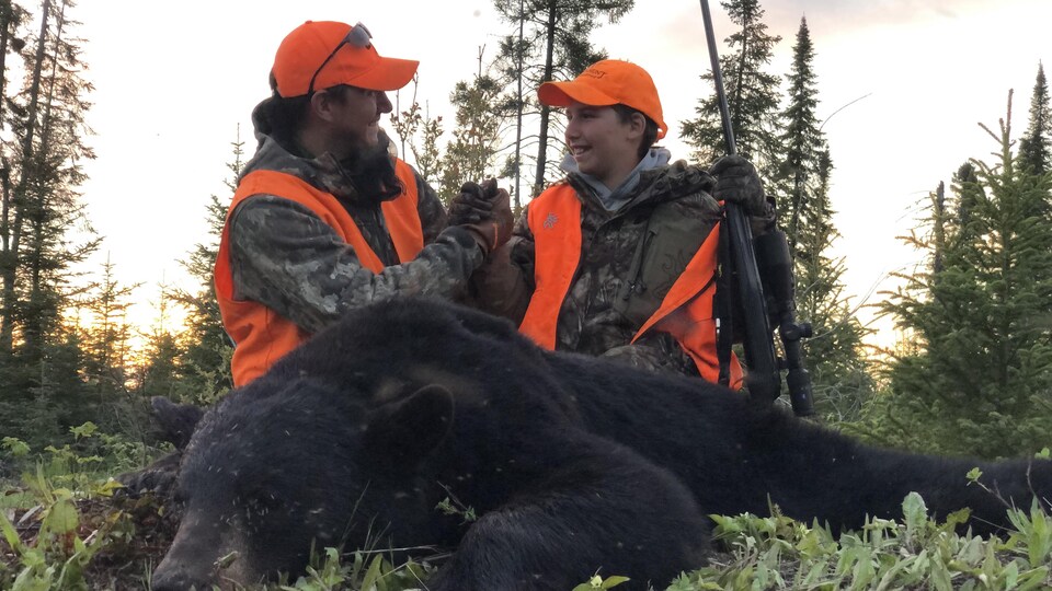 Un père et son fils vêtus de vêtements de camouflage et de vestes oranges se regardent et sourient, visiblement heureux d'avoir abattu l'ours noir qui se trouve juste devant eux.