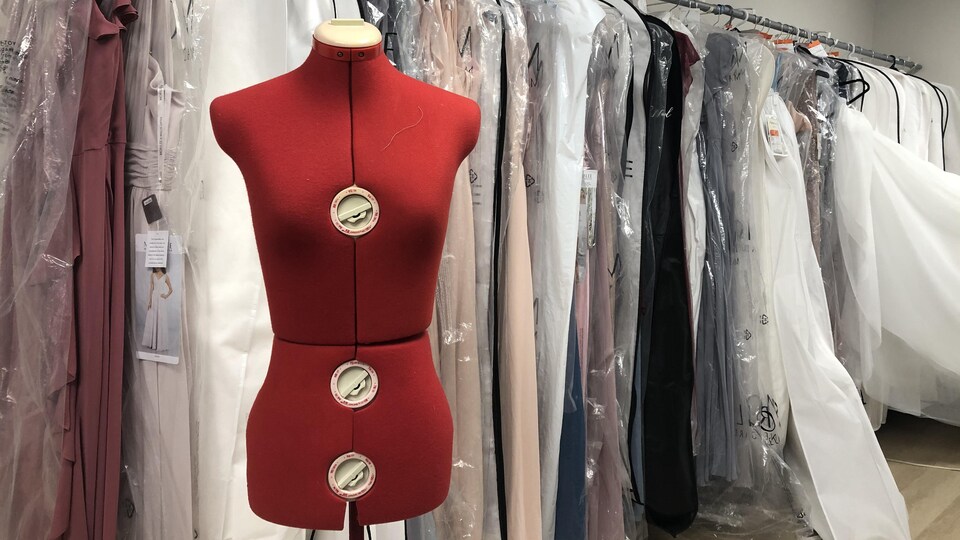 Un mannequin de couture devant plusieurs robes protégées d'un plastique, suspendues sur une longue tringle.