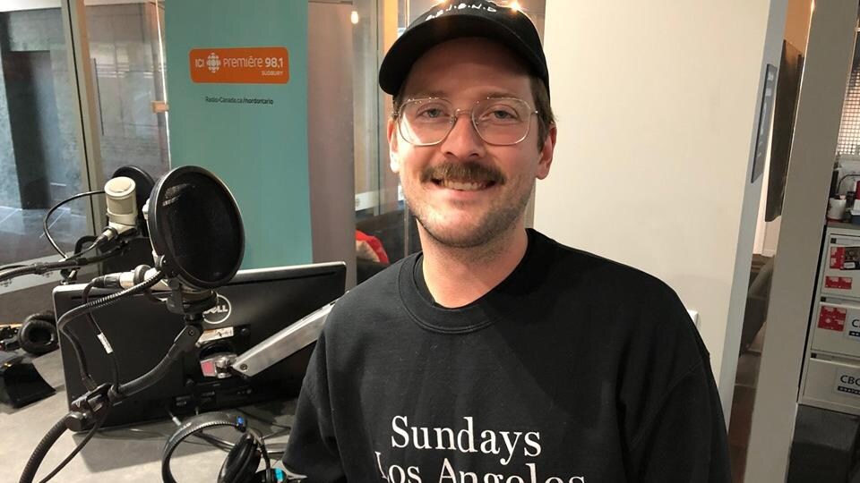 Frank Bach, graphiste principal de l'application mobile Headspace, dans le studio de Sudbury sourit à la caméra vêtue d'un chandail noir et d'une casquette noire