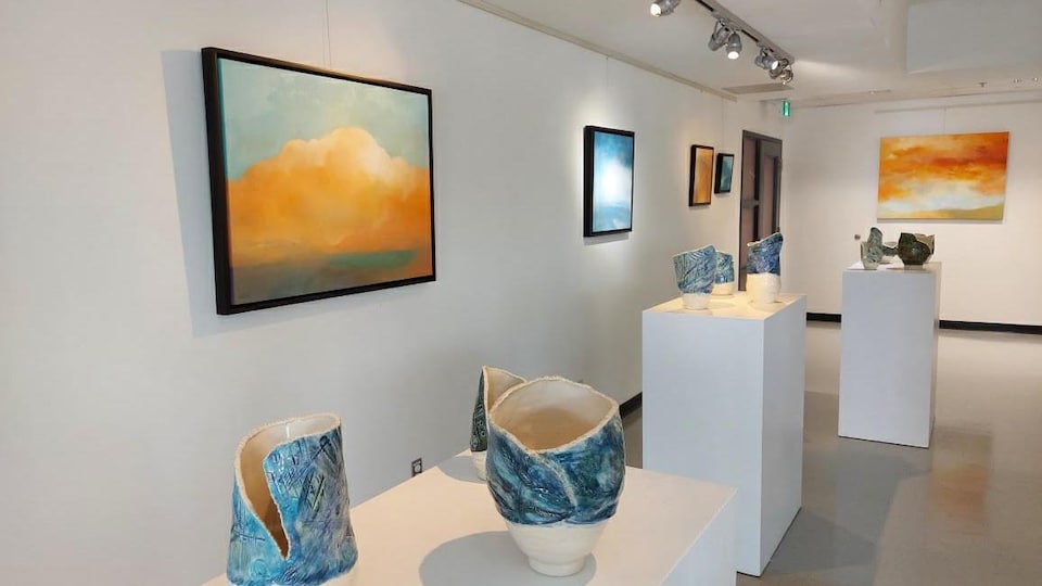 Une galerie d'art où sont accrochés des tableaux de cieux ennuagés; des pots en céramique sont exposés sur des socles