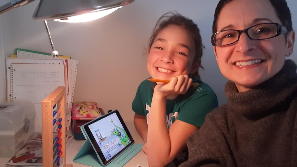 Une mère et sa jeune fille sont à la table avec du matériel électronique, du papier et des crayons.