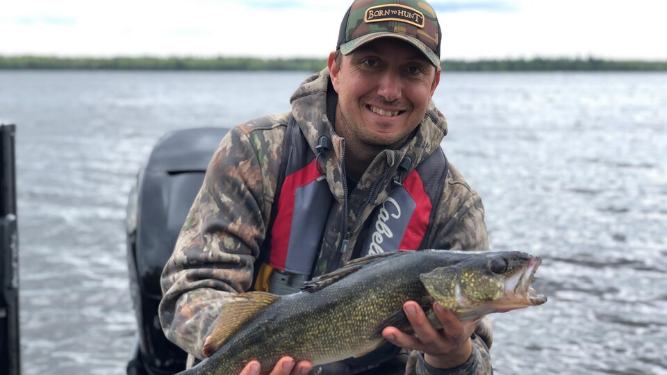 Un homme sourit à la caméra en tenant un gros poisson qu'il vient de capter. Il est dans un bateau sur un lac agité. 