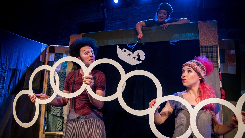 Trois comédiens manipulent des anneaux et la marionnette d'un navire devant une scène improvisée.