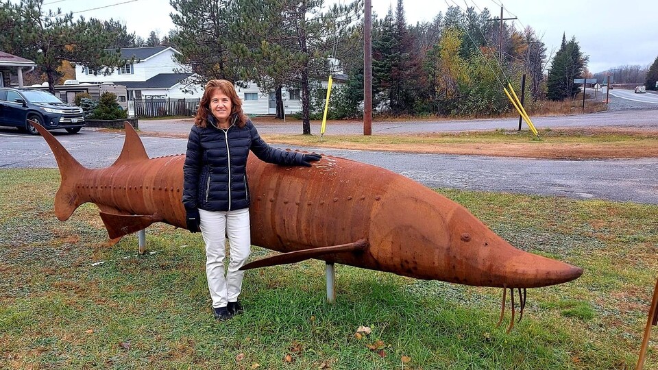 Gayle Primeau devant la sculpture en métal d’un immense esturgeon.