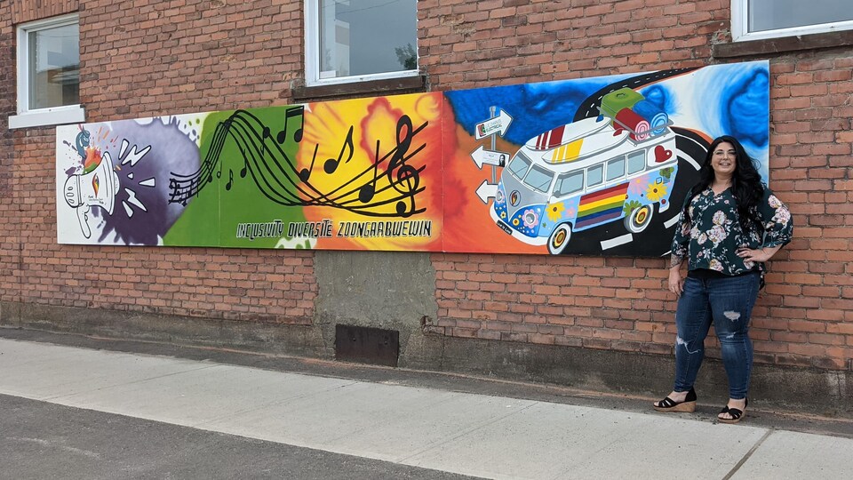 Tracy DeCaen pose devant sa murale; trois panneaux en format paysage accrochés sur un mur de brique