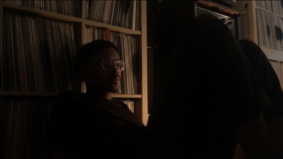Akeem Oh assis dans une salle sombre, une grande collection de disques vinyle derrière lui