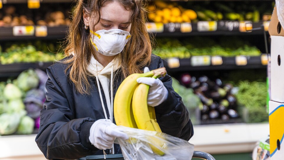 Une femme portant un masque et des gants place des bananes dans un sac en plastique.