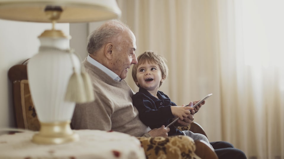 Un vieille homme tient assis sur ses cuisses un enfant qui le regarde en souriant.  