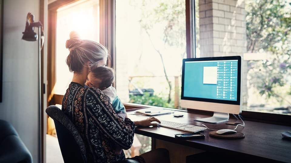 Une femme avec son bébé dans les bras tandis qu'elle travaille sur un ordinateur.