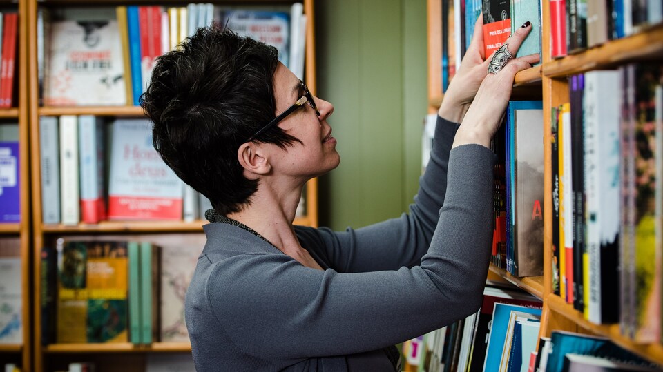 On voit une femme qui se trouve dans une bibliothèque. Elle manipule quelques livres dans une étagère.