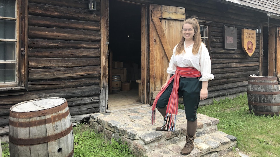 Une femme portant la ceinture fléchée se tient devant un bâtiment d'époque en bois.