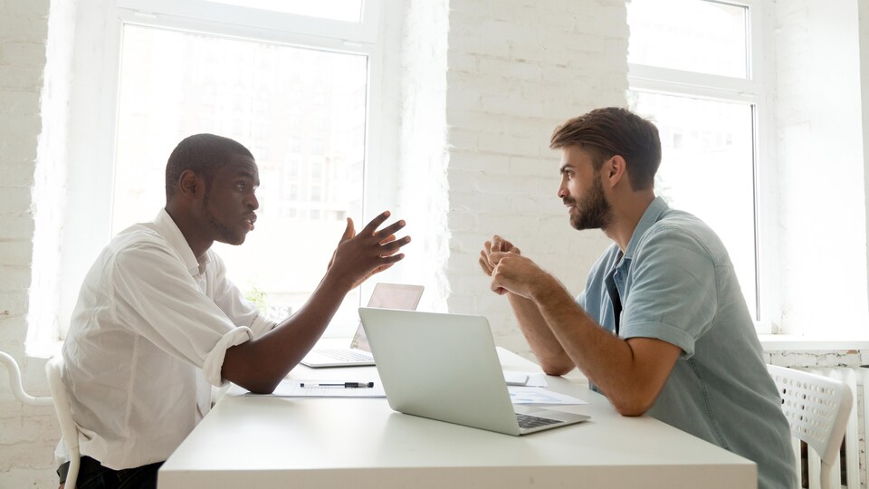 Deux hommes sont assis de chaque côté d'un table sur laquelle est posée deux ordinateurs.