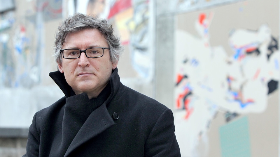 Assis vêtu d'un manteau noir, il porte des lunettes noires et des cheveux grisonnants.
