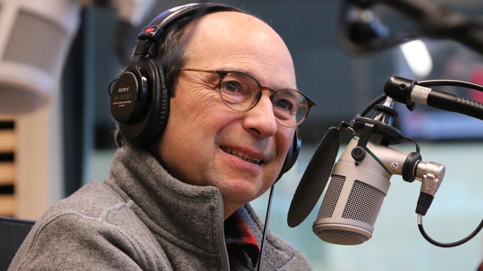 Un homme, écouteurs sur la tête, sourit devant un micro dans un studio de radio. 