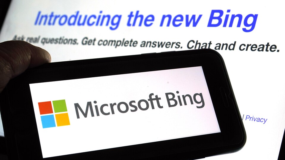 Le logo de Microsoft Bing est présenté en plein écran sur un téléphone cellulaire devant l'écran d'un moniteur qui affiche la page du site web de Microsoft, où on lit un texte au sujet de son nouveau robot conversationnel.