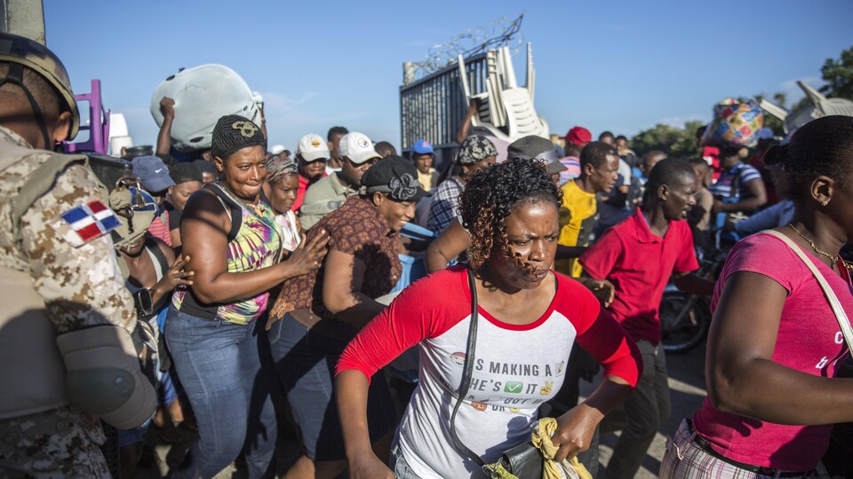 Groupe de citoyens haïtiens s'empressant de traverser la frontière, munis de marchandises, sous la supervision armée d'un soldat