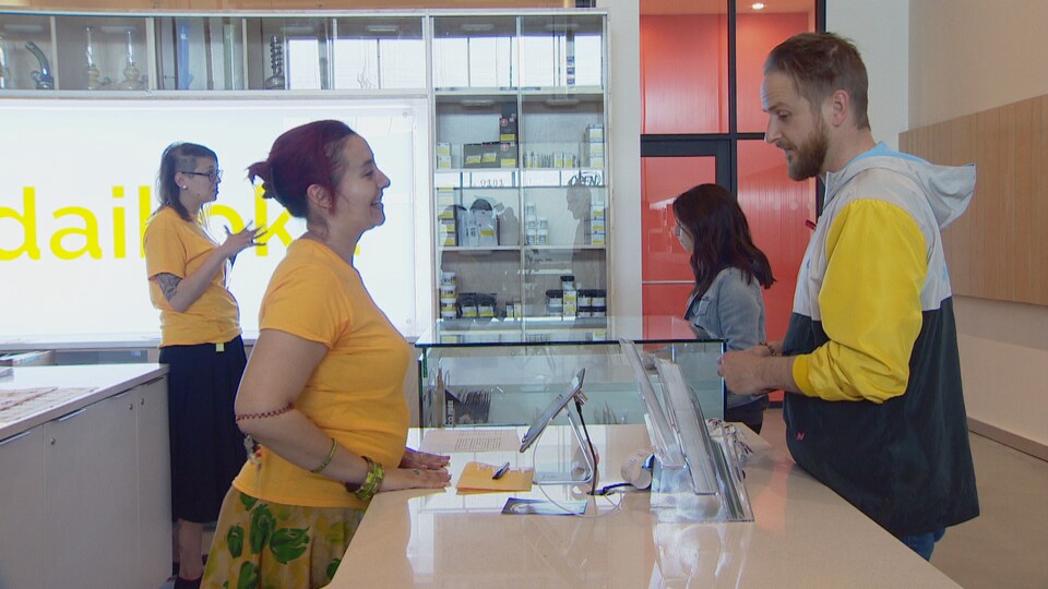 Dans une boutique. Mireille Tessier et une employée portent des gilets jaunes. Elles sont en train de répondre à des clients en souriant et en gesticulant.