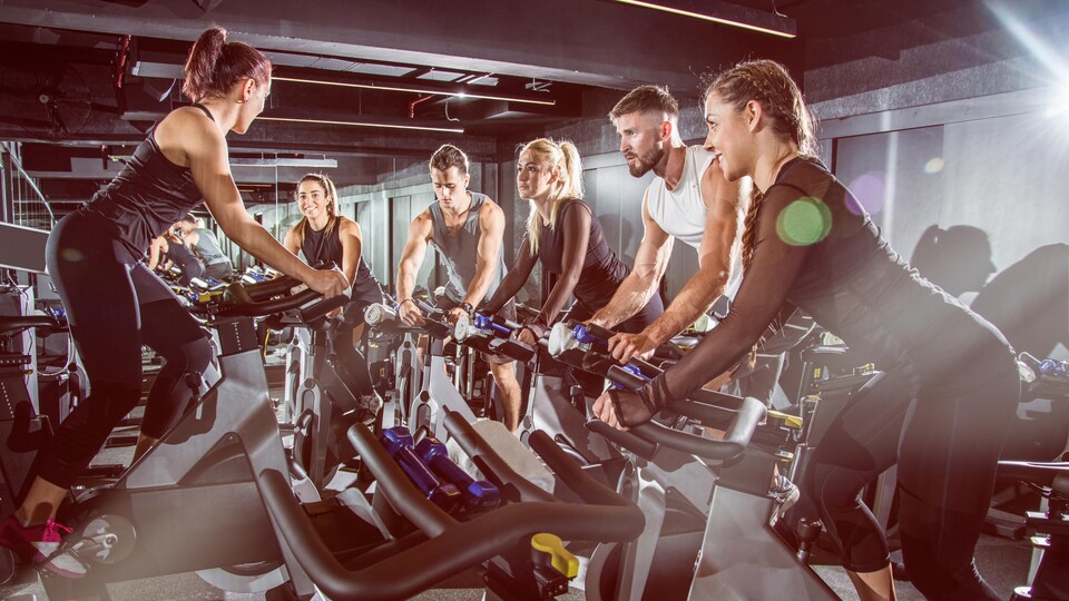 Des hommes et des femmes, jeunes et musclés, font du vélo stationnaire en salle d'entraînement, sous une lumière naturelle radieuse.