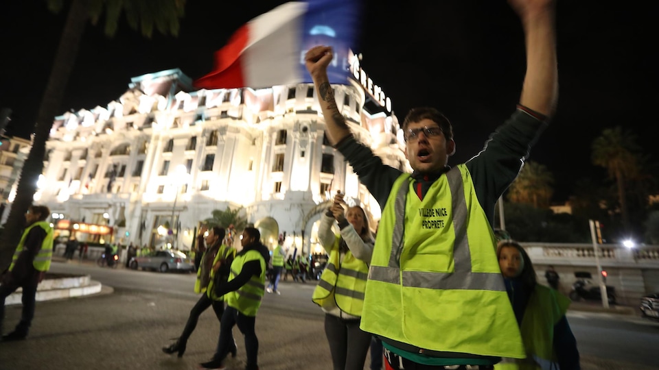 Un homme avec un gilet jaune lève les bras. Derrière lui, une femme avec un gilet jaune tient un drapeau français, et d'autres gens marchent dans la rue.