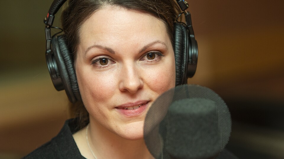 La comédienne derrière un micro dans un studio de radio. Elle porte des écouteurs.
