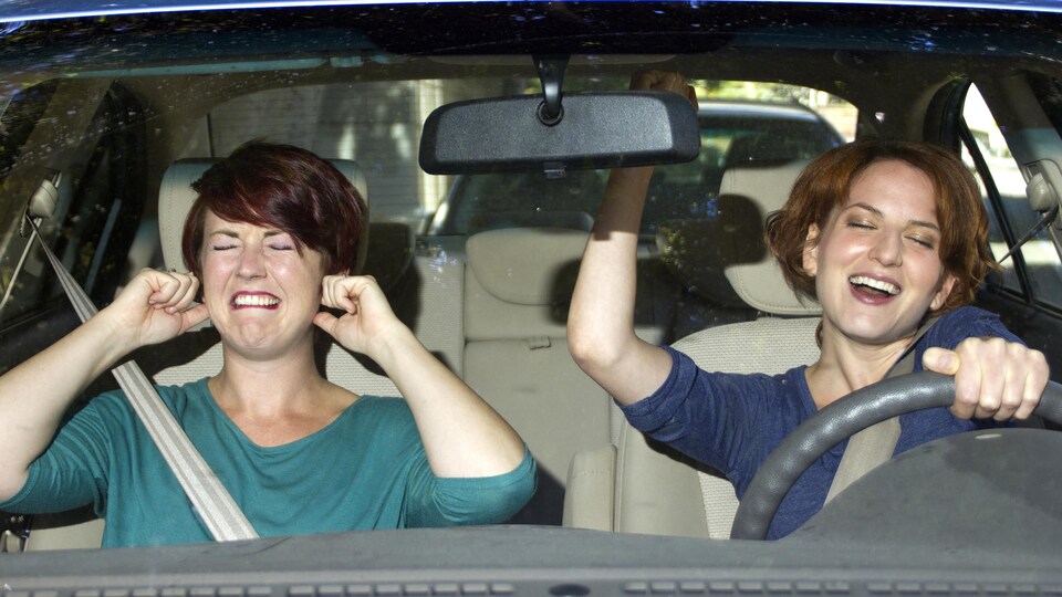 Une femme chante au volant pendant que sa passagère se bouche les oreilles.