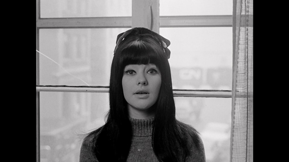 Photo en noir et blanc d'une femme, devant une fenêtre, qui regarde la caméra.