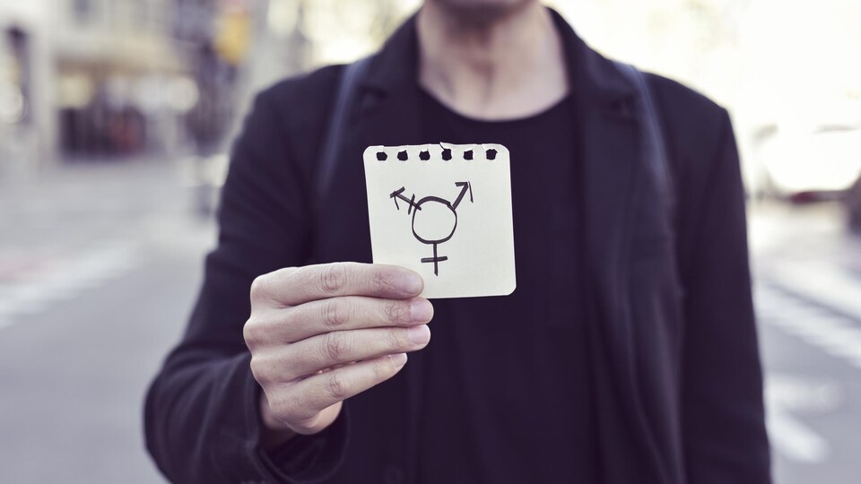Une personne tient un papier sur lequel est dessiné le symbole transgenre.