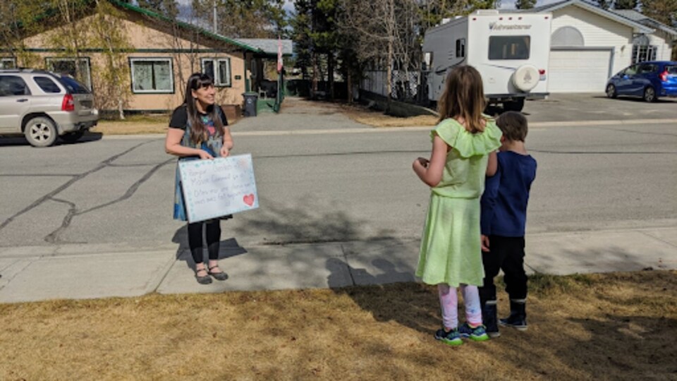 Une femme avec un tableau parle à deux enfants devant une maison