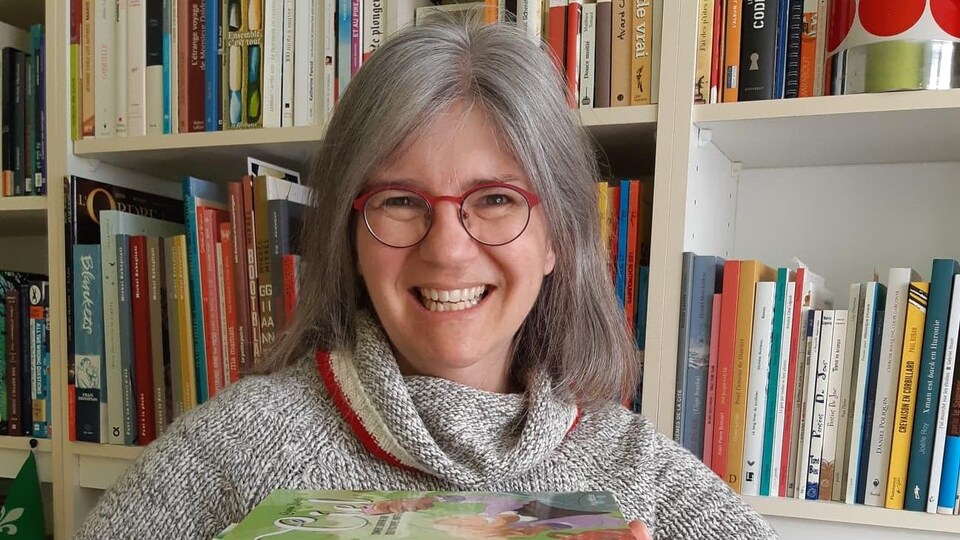 Mireille Messier tiens plusieurs livre devant sa bibliothèque